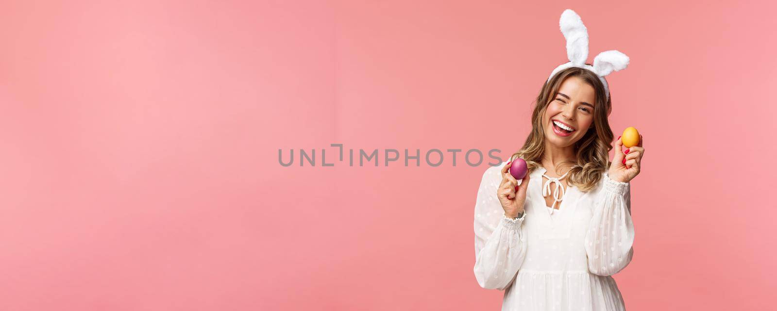 Holidays, spring and party concept. Cheerful good-looking blond woman celebrating Easter day in rabbit ears, holding two painted eggs and wink camera, smiling happily, pink background by Benzoix