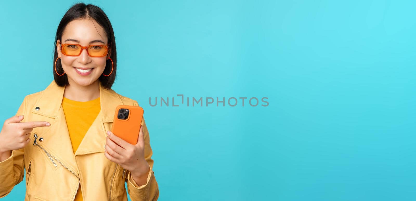 Young stylish asian female model in trendy sunglasses, spring outfit, showing mobile phone, pointing at smartphone and smiling, standing over blue background.