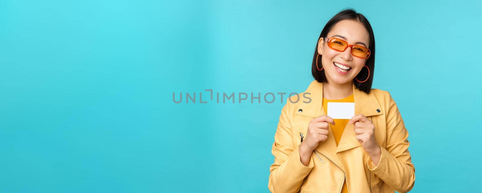 Stylish young asian woman in sunglasses, showing credit card and smiling, recommending bank, contactless payment or discounts in store, standing over blue background by Benzoix