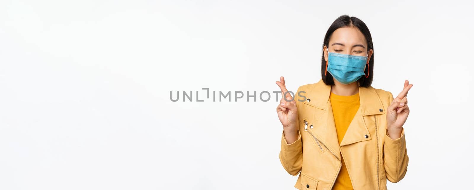 Hopeful asian girl in medical face mask, cross fingers, making wish, hoping, praying for smth, standing with anticipation over white studio background.