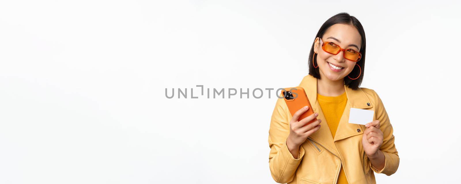 Online shopping and delivery concept. Happy korean girl in stylish clothes, holding credit card and smartphone, laughing and smiling, standing over white background.