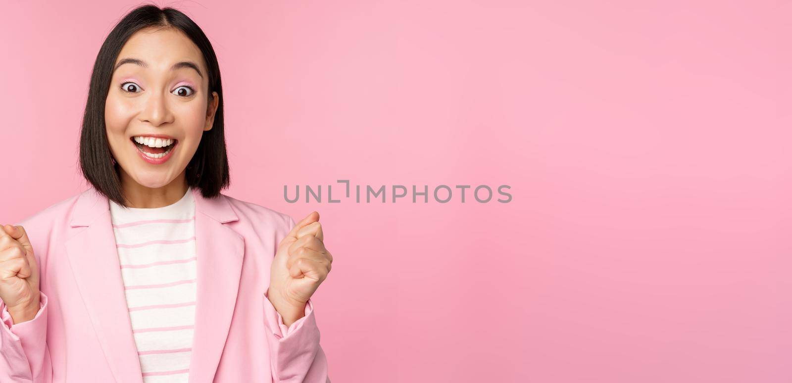 Enthusiastic saleswoman, asian corporate woman say yes, achieve goal and celebrating, triumphing, looking with rejoice and smiling, standing over pink background by Benzoix