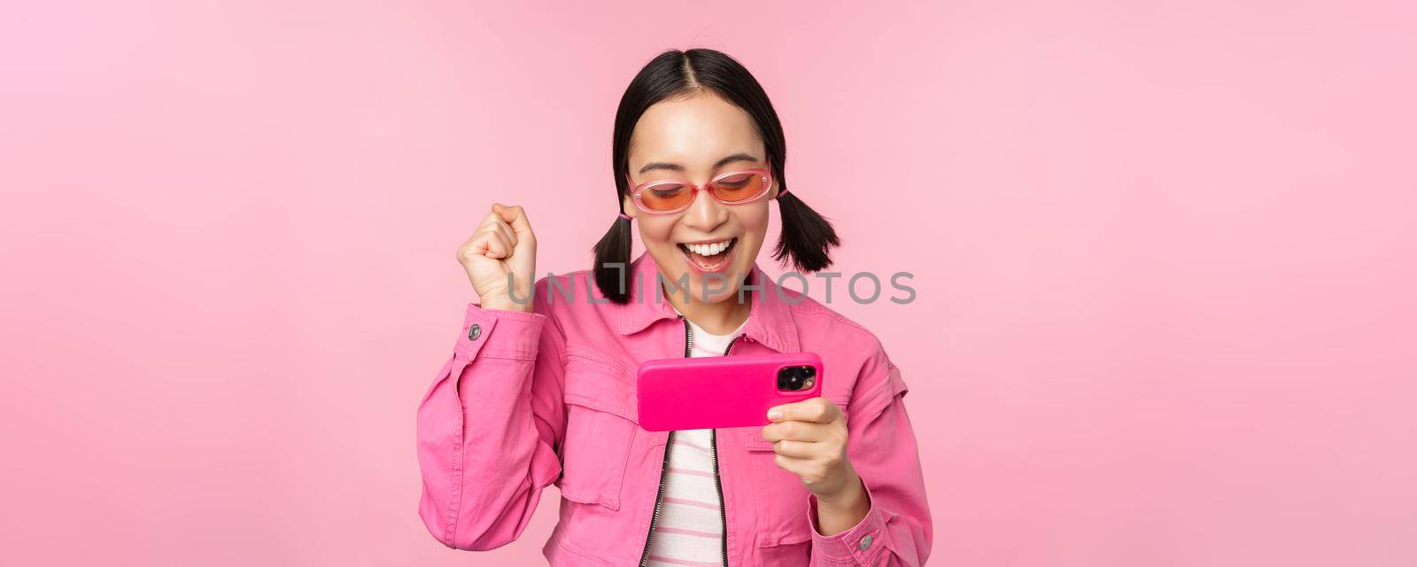 Happy smiling korean girl winning on mobile phone, looking at horizontal smartphone screen and rejoicing, achieve goal, celebrating, standing over pink background by Benzoix