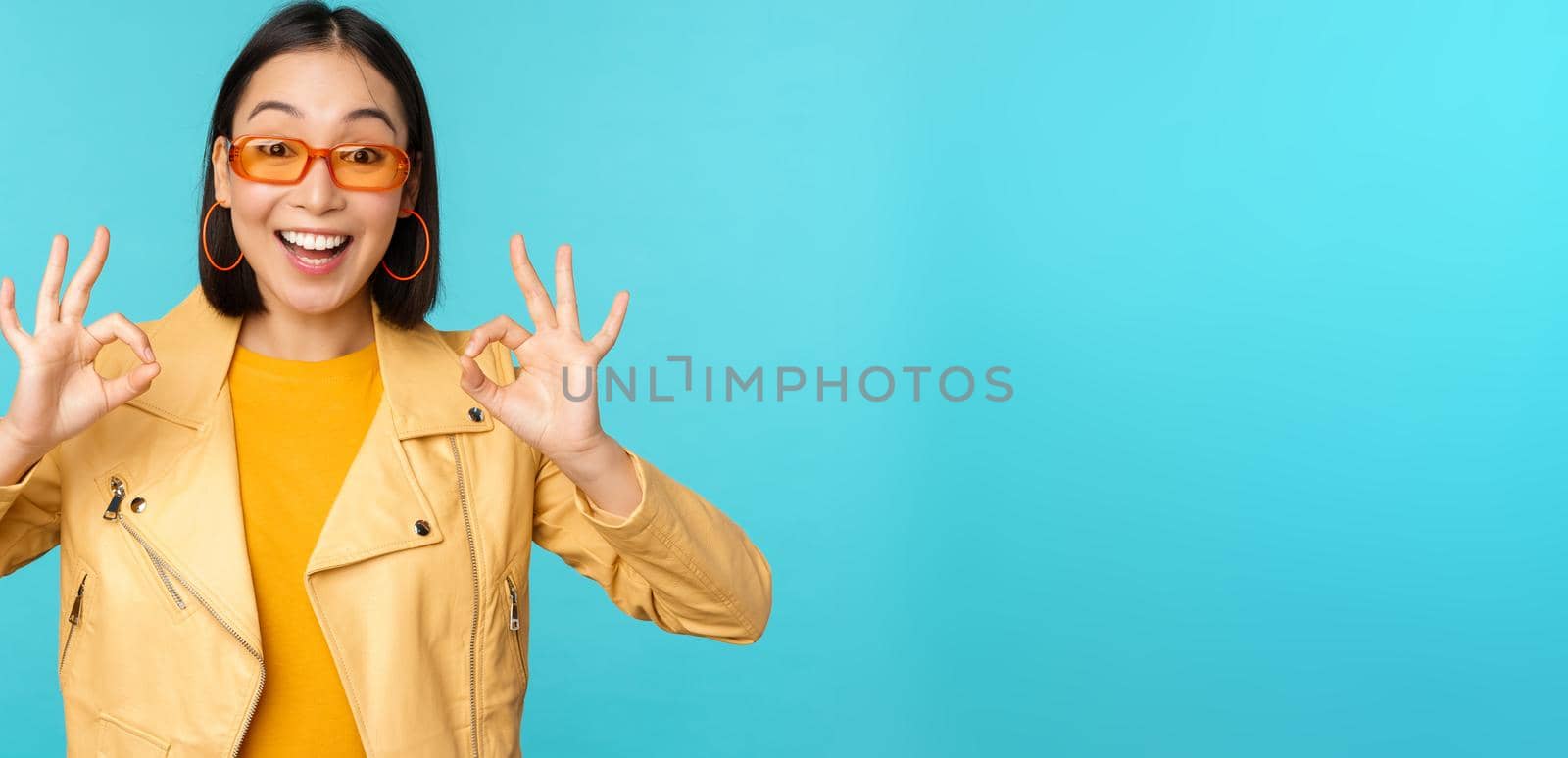 Stylish beautiful asian woman in sunglasses, smiling amazed, showing okay, ok sign, recommending smth, supports excellent choice, standing over blue background.