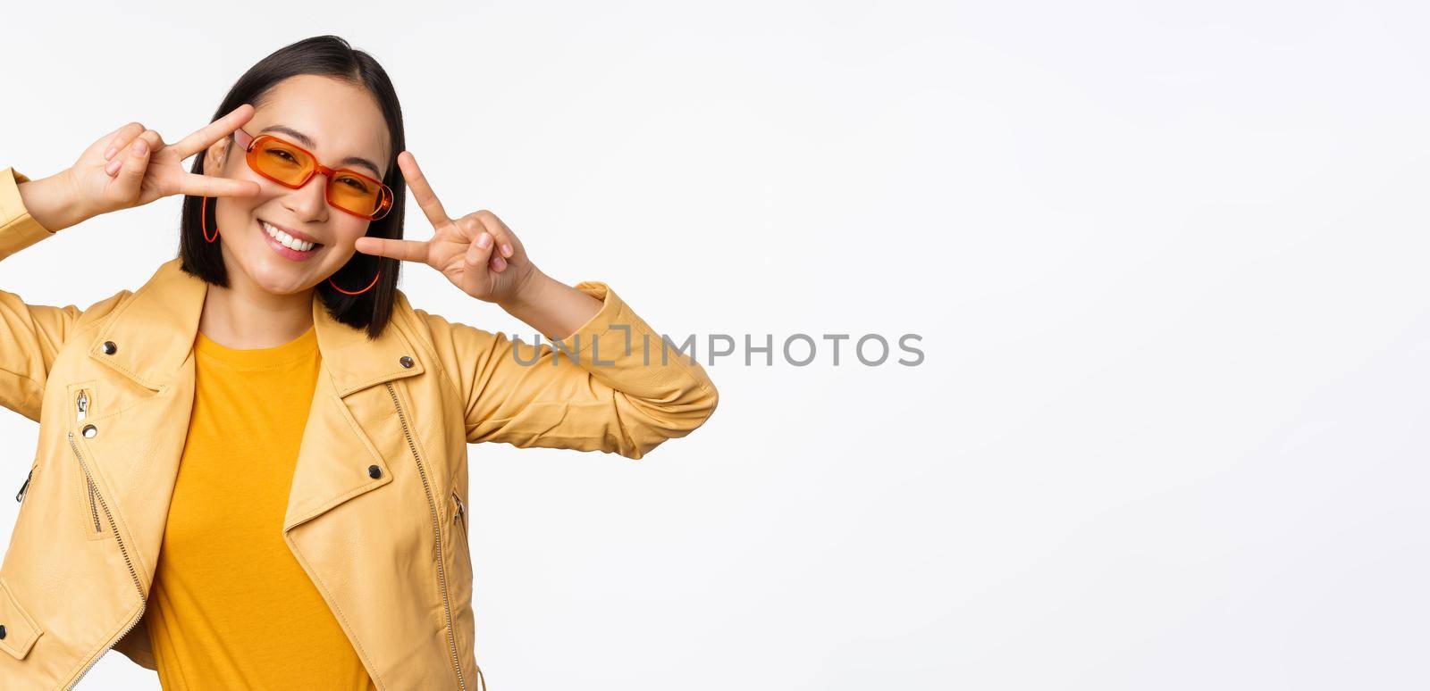 Portrait of stylish asian modern girl, wearing sunglasses and yellow jacket, showing peace, v-sign gesture, standing over white background, happy smiling face.