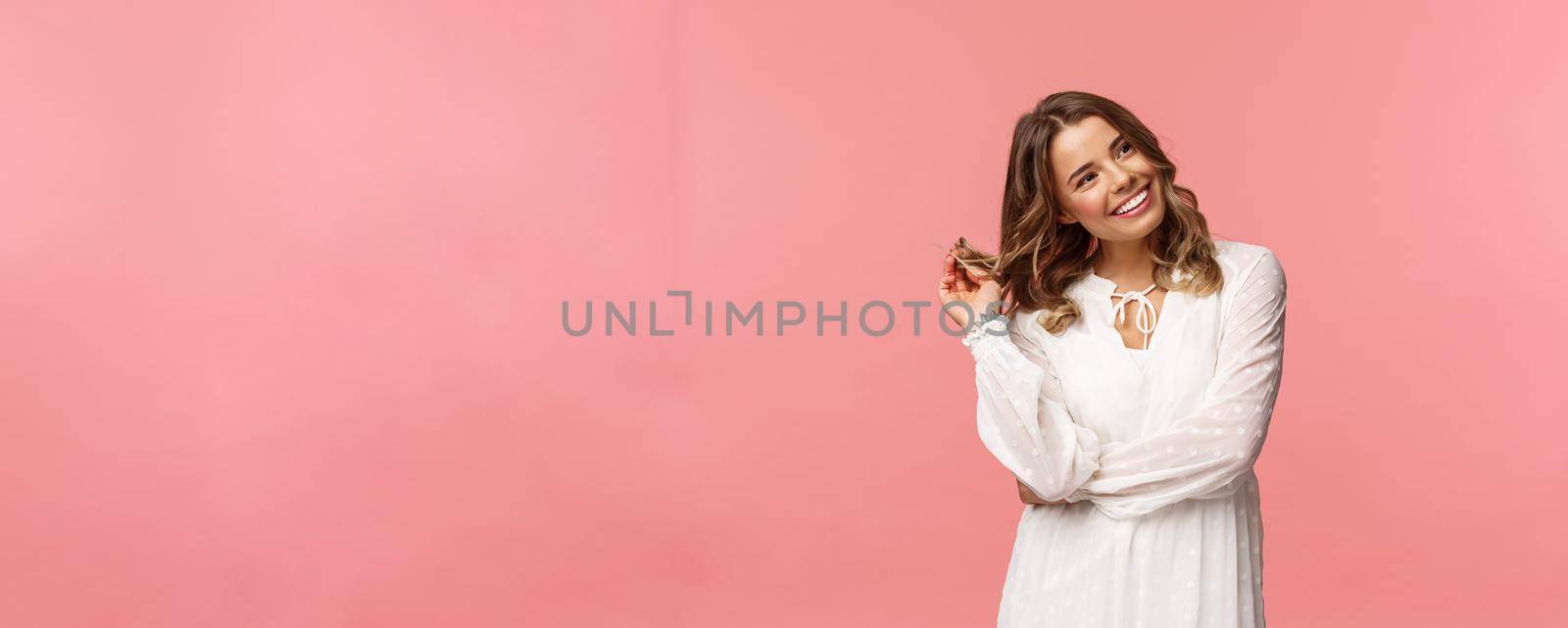 Feminine, beautiful and coquettish young blond woman in stylish white sping dress, giggle silly look away at right side, rolling curl on hair strand flirting with someone over pink background.