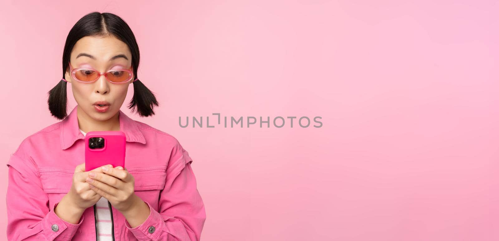 Portrait of asian girl in sunglasses using smartphone. Woman looking at mobile phone, browsing in app, standing over pink background.
