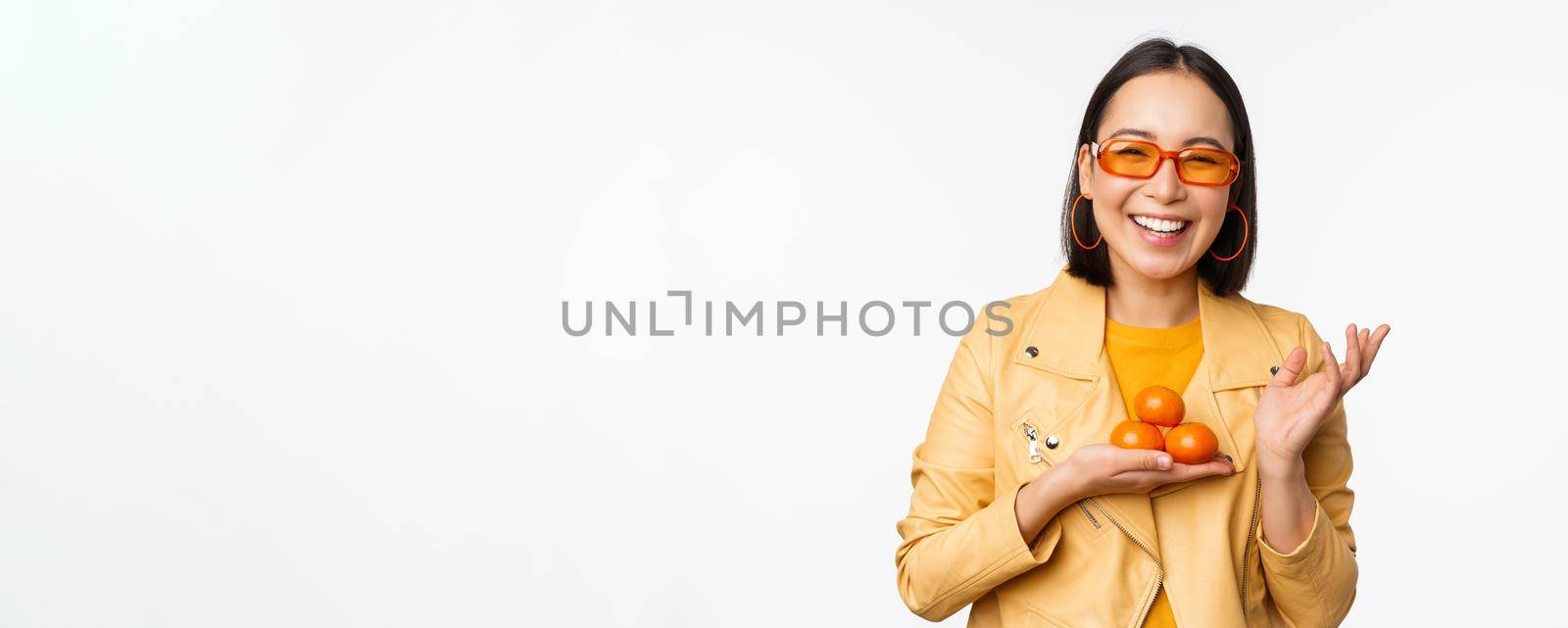 Stylish happy asian girl in sunglasses holding tangerines and smiling, posing against white background. Copy space