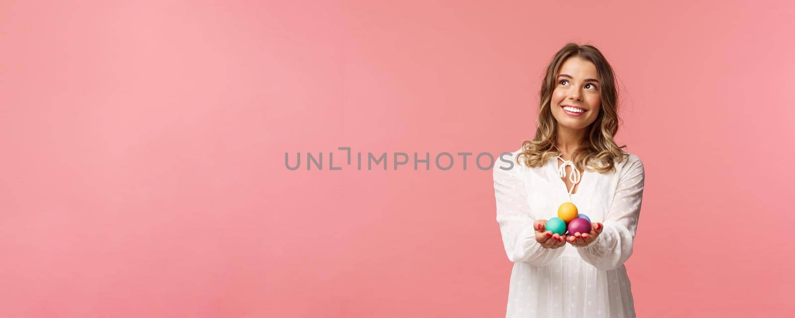 Holidays, spring and party concept. Portrait of dreamy good-looking blond girl in white dress, holding painted eggs, celebrating Easter, orthodox holy day, smiling and looking away.