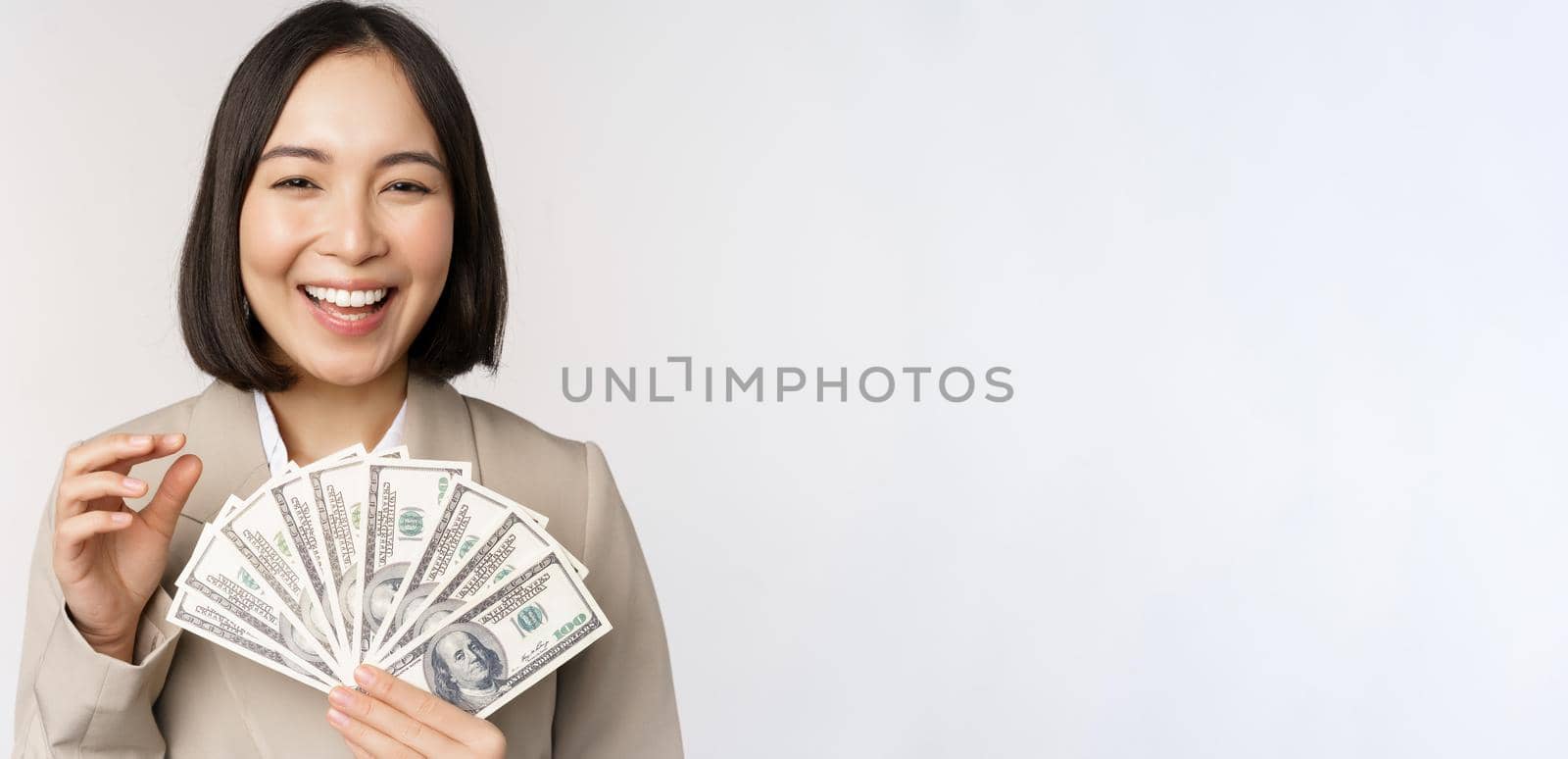 Image of successful businesswoman holding money. Asian corporate woman with cash dollars, smiling and laughing, standing over white background.