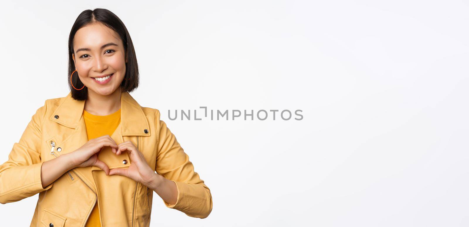 Image of beautiful asian girl showing heart, love sign, express care and romantic feelings, standing over white studio background.