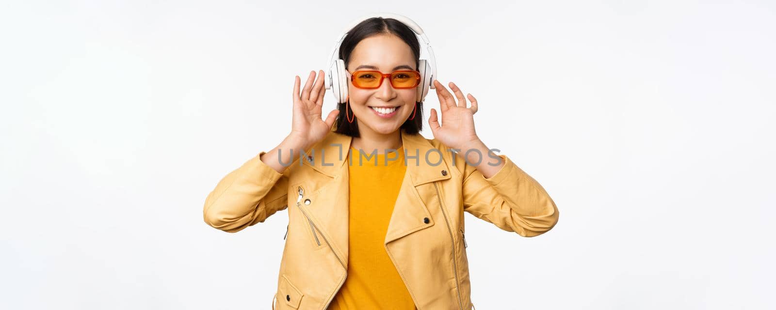 Beautiful asian girl, laughing happy, listening music in headphones, standing over white background. Copy space