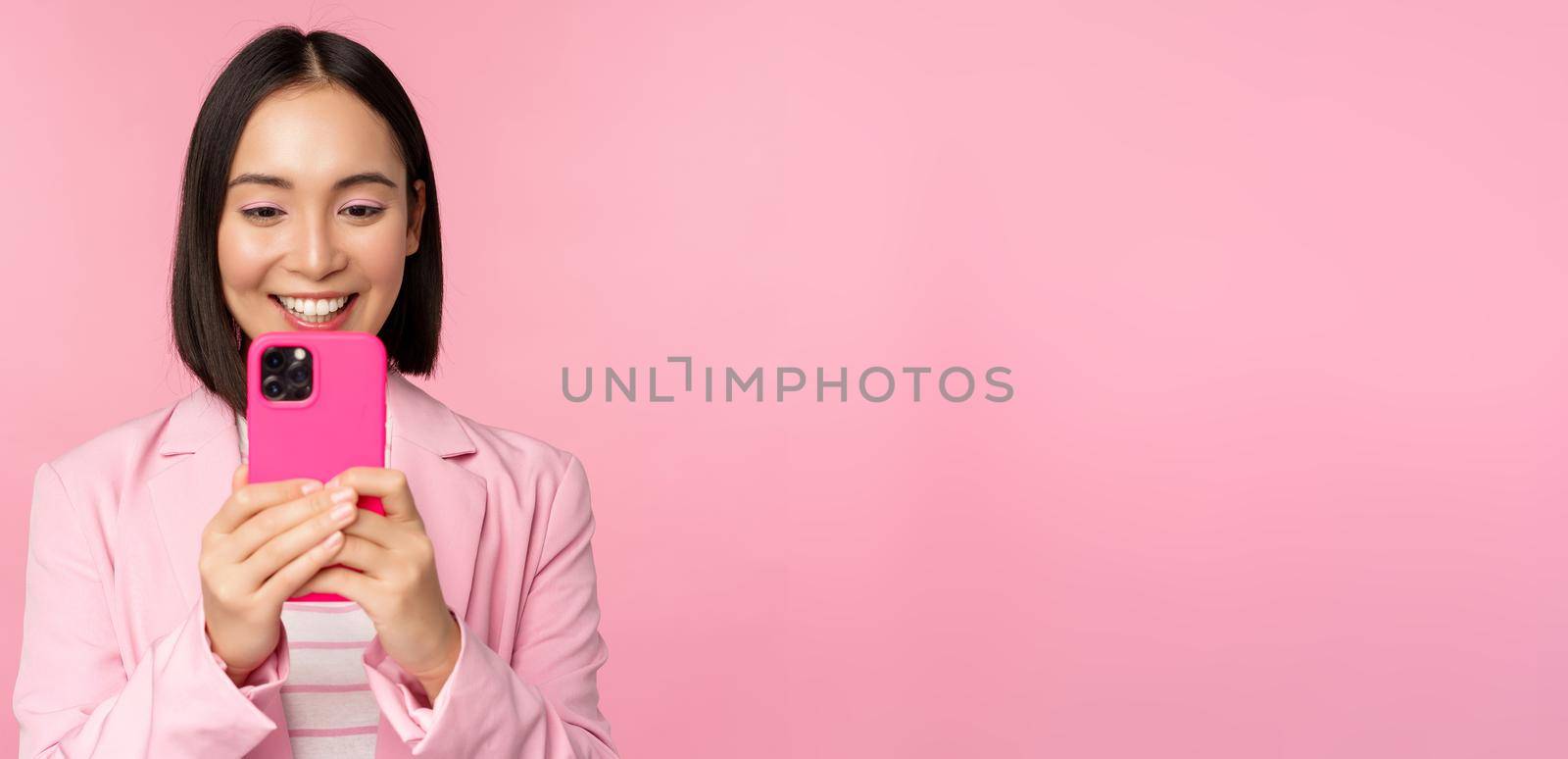 Image of smiling asian corporate woman in suit looking, watching on smartphone app, using mobile phone application, standing over pink background by Benzoix