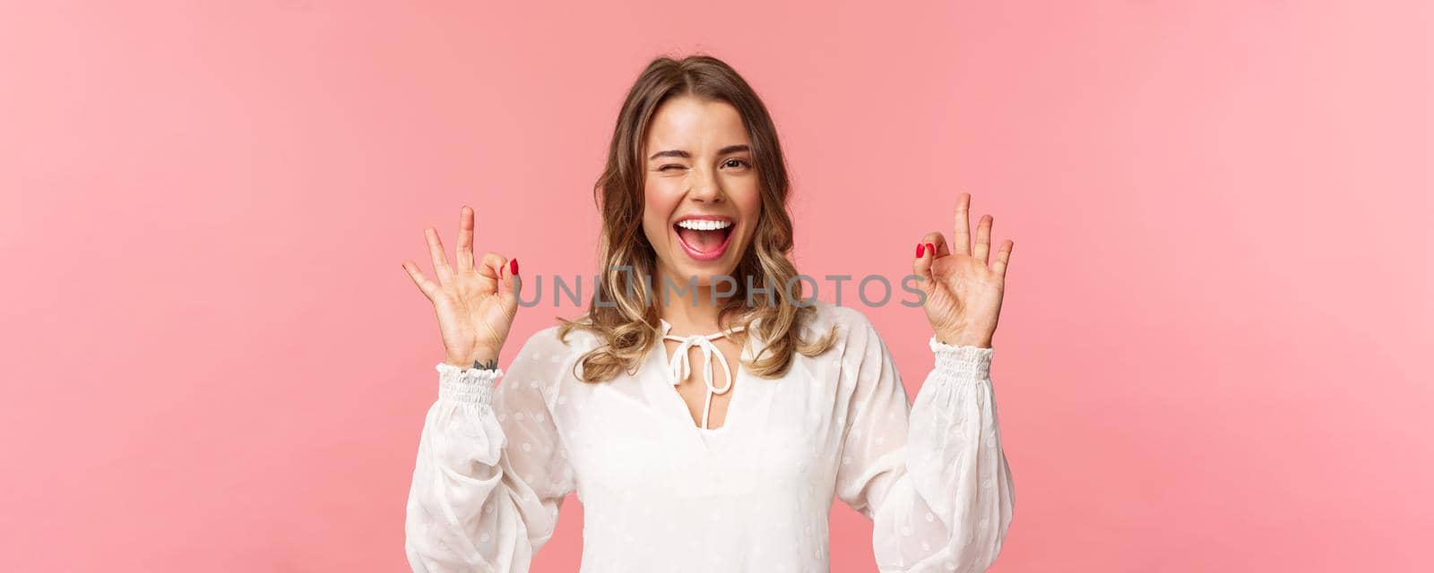 Close-up portrait of beautiful blond girl in white dress guarantee you will enjoy this event, show okay sign, confirm everything excellent, wink and smiling carefree, standing pink background by Benzoix