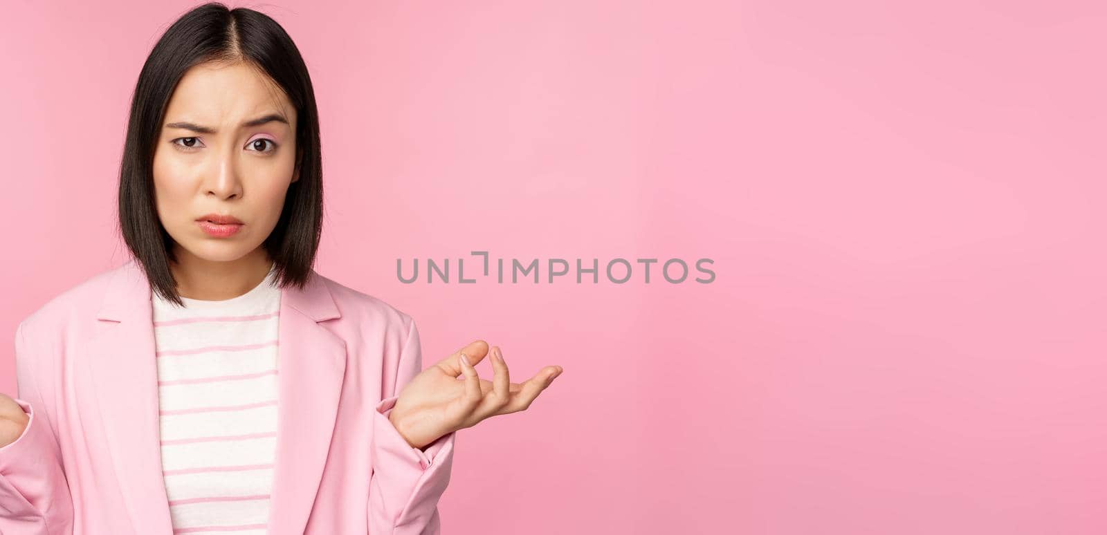 Portrait of young asian business woman, saleswoman shrugging shoulders and looking confused, clueless of smth, standing over pink background.