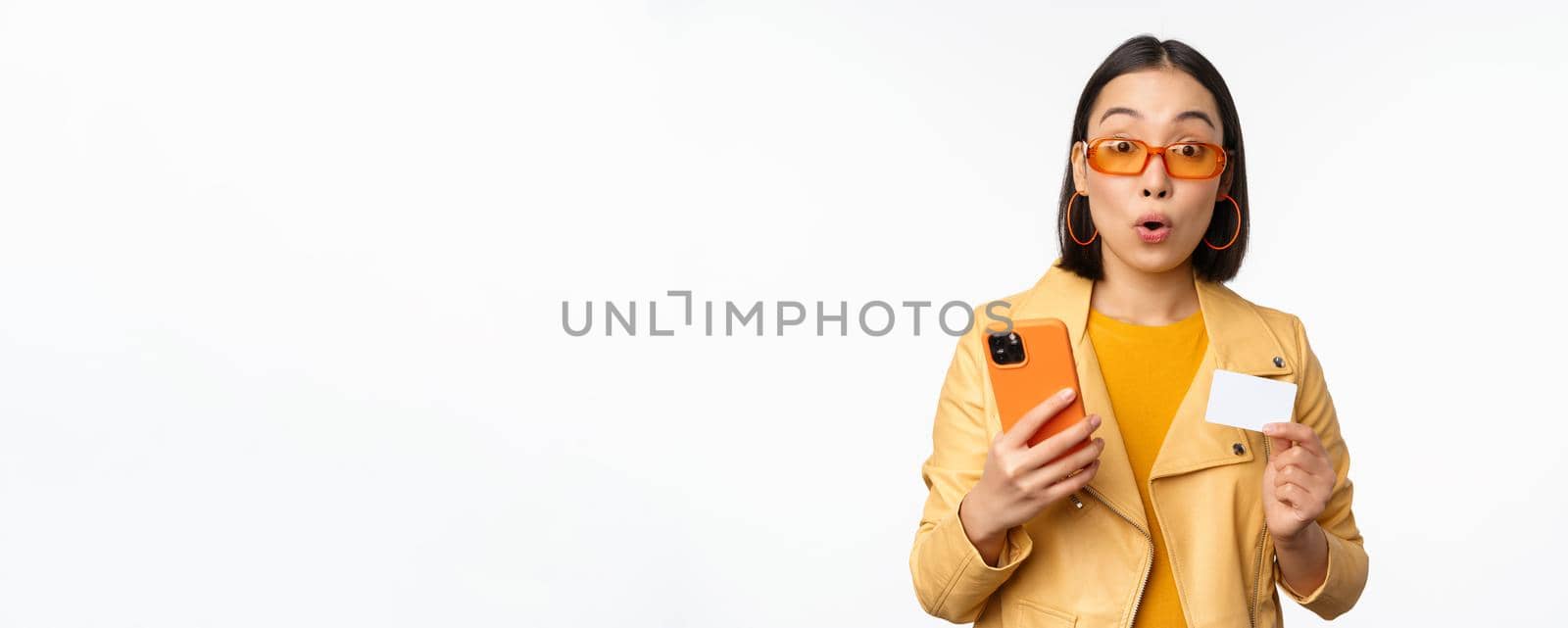 Online shopping. Stylish asian female model in sunglasses, holding credit card and mobile phone, smiling happy, standing over white background by Benzoix