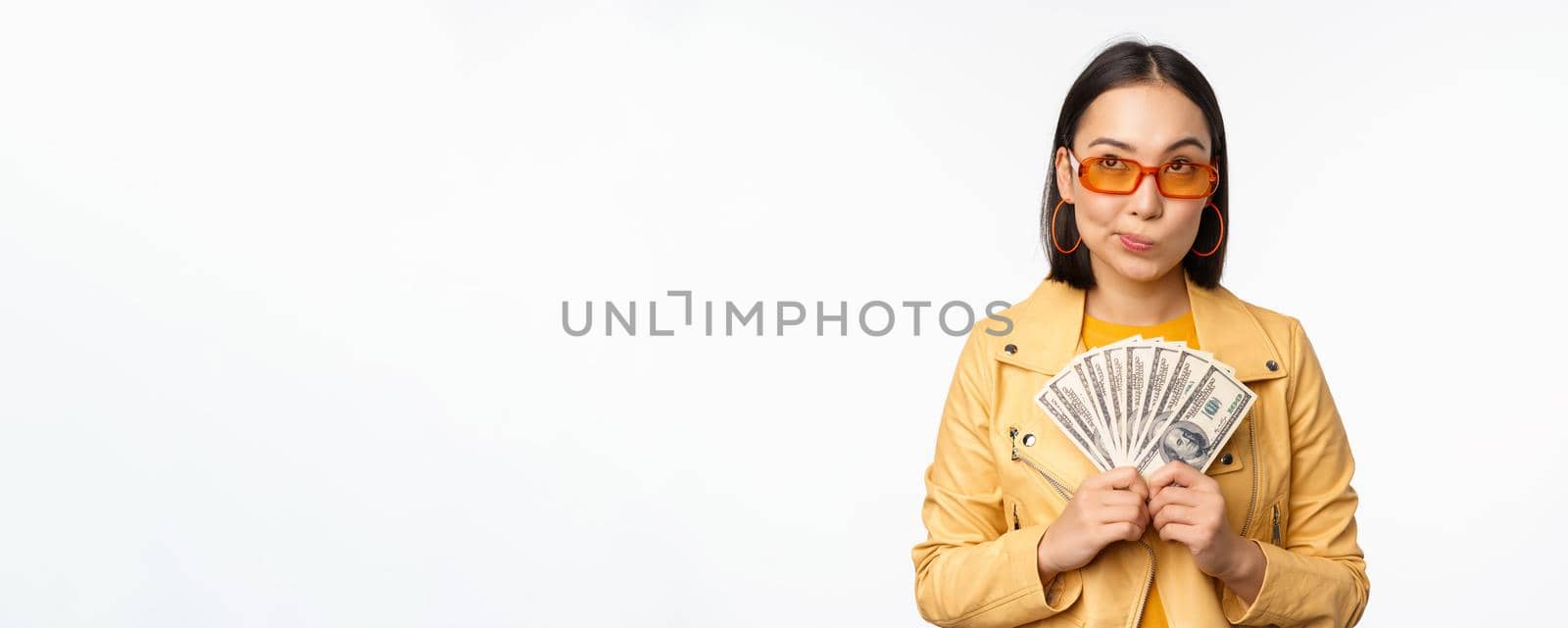 Microcredit and money concept. Stylish asian young woman in sunglasses, laughing happy, holding dollars cash, standing over white background.