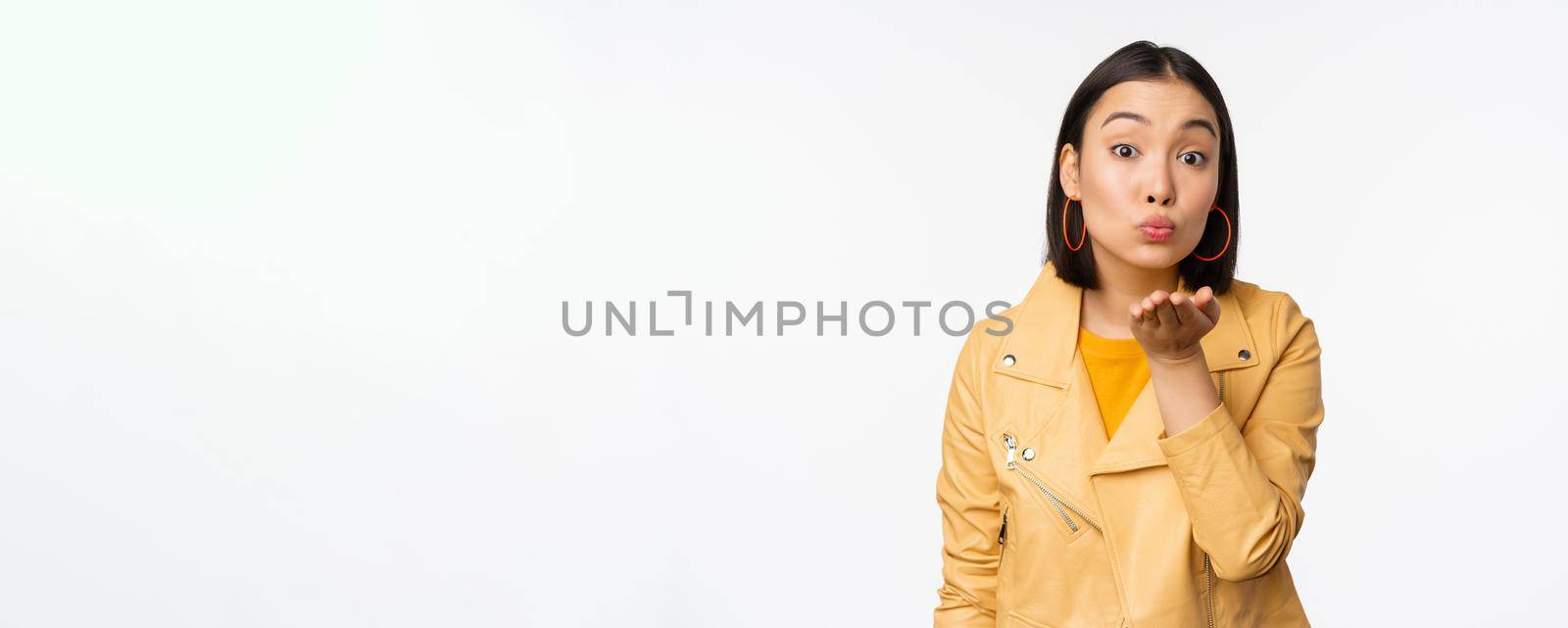 Cute korean stylish girl, sending air kiss, blowing mwah and looking silly romantic at camera, standing over white background. Copy space