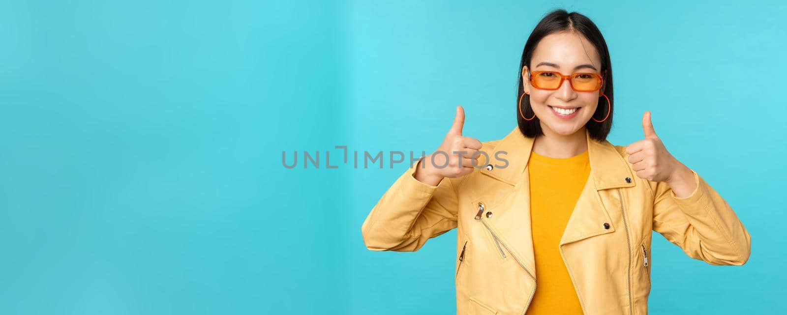 Portrait of modern young asian chinese woman in sunglasses, wears trendy outerwear, showing thumbs up and smiling pleased, recommending smth, blue background.