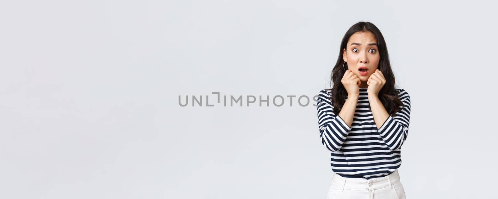 Lifestyle, people emotions and casual concept. Scared worried asian girl in striped shirt, gasping looking alarmed, got in trouble, find out shocking truth, standing white background.