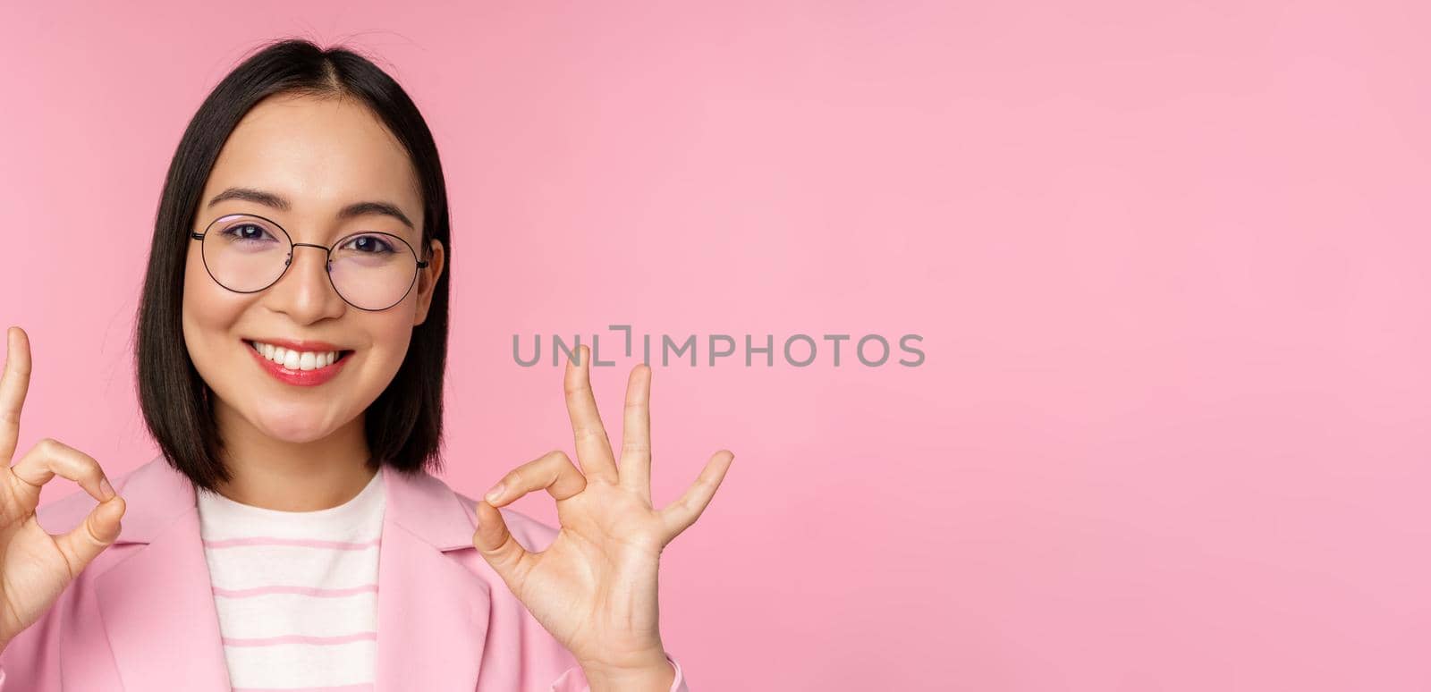 Close up portrait of impressed corporate woman, asian business lady in glasses, showing okay sign, looking amazed at camera, recommending, pink background by Benzoix