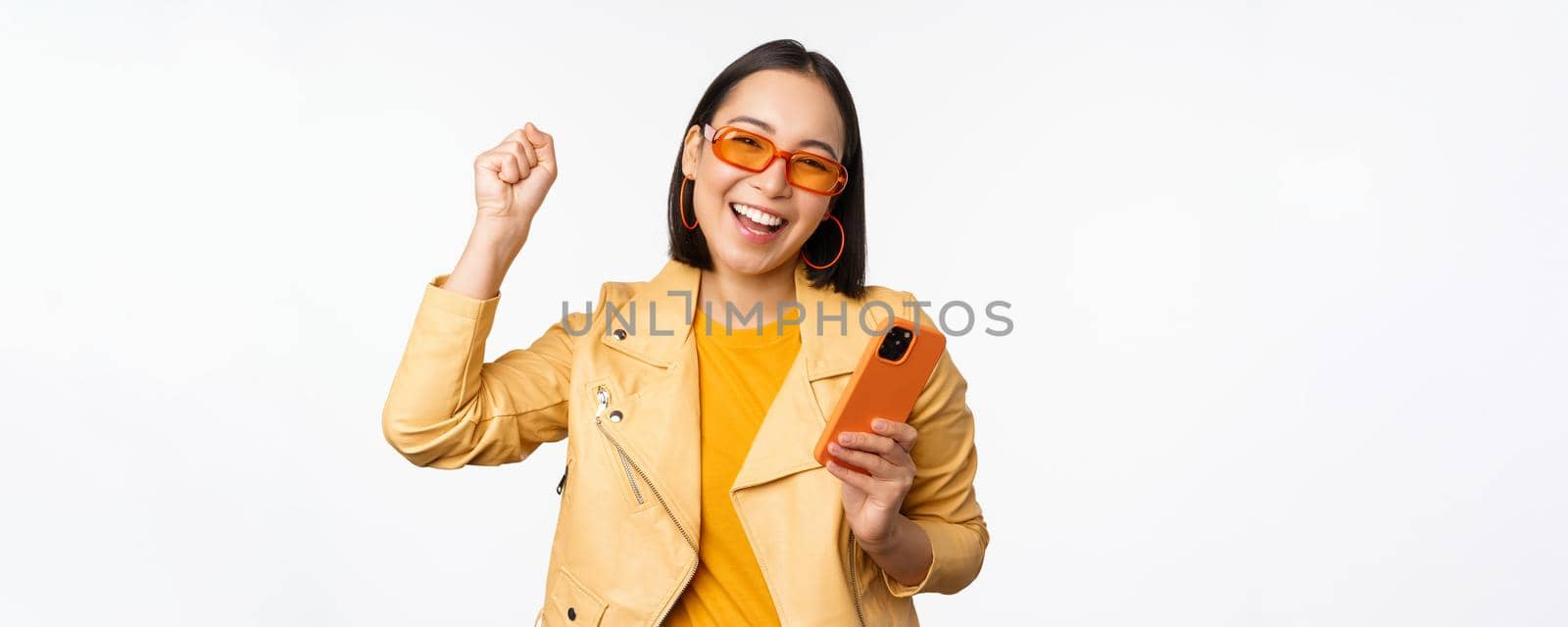 Enthusiastic korean girl in sunglasses, holding smartphone, celebrating and dancing, laughing happy with mobile phone, standing over white background.