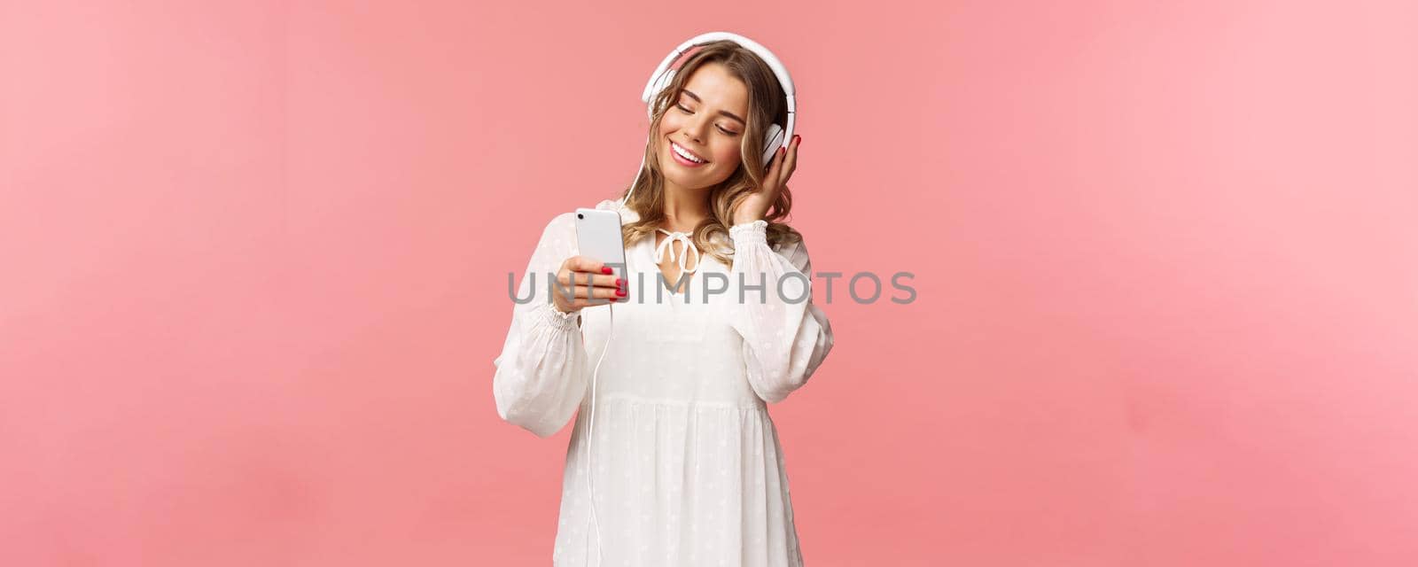 Portrait of dreamy cute and tender blond girl in white dress, holding smartphone, listen music in headphones, smiling at display mobile phone as picking playlist for spring mood, pink background.