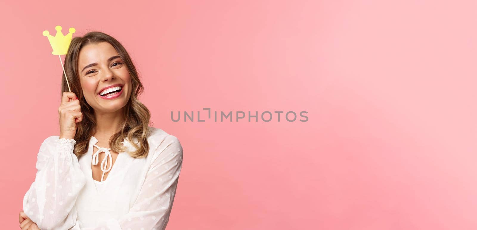 Spring, happiness and celebration concept. Close-up portrait of charming smiling, lovely blond girl holding small queen crown on stick, laughing joyfully, feel empowered and happy, pink background by Benzoix
