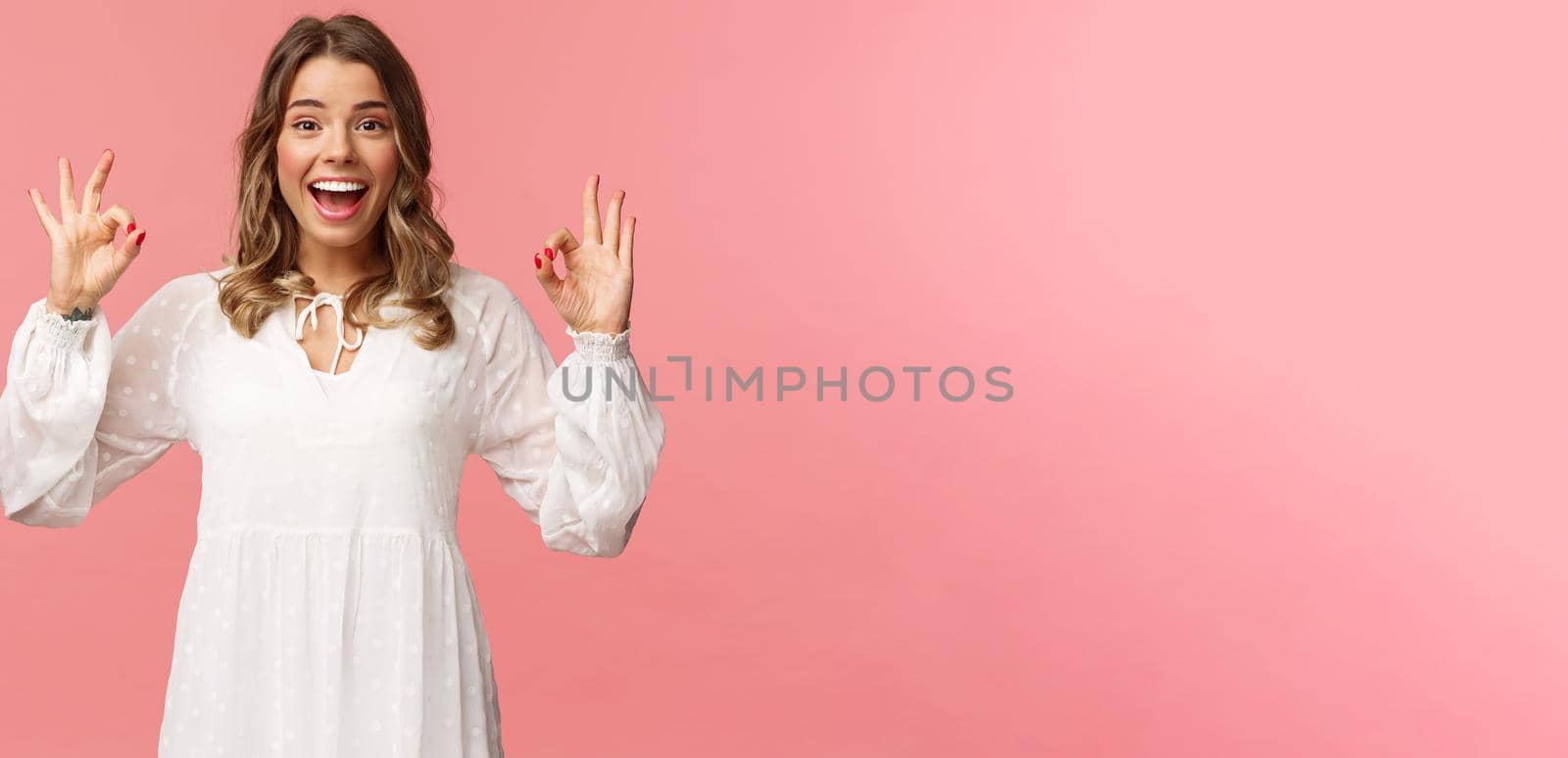 Portrait of happy and pleased good-looking blond woman in white dress, recommend great product, leave positive review, show okay signs and smiling satisfied, standing pink background.