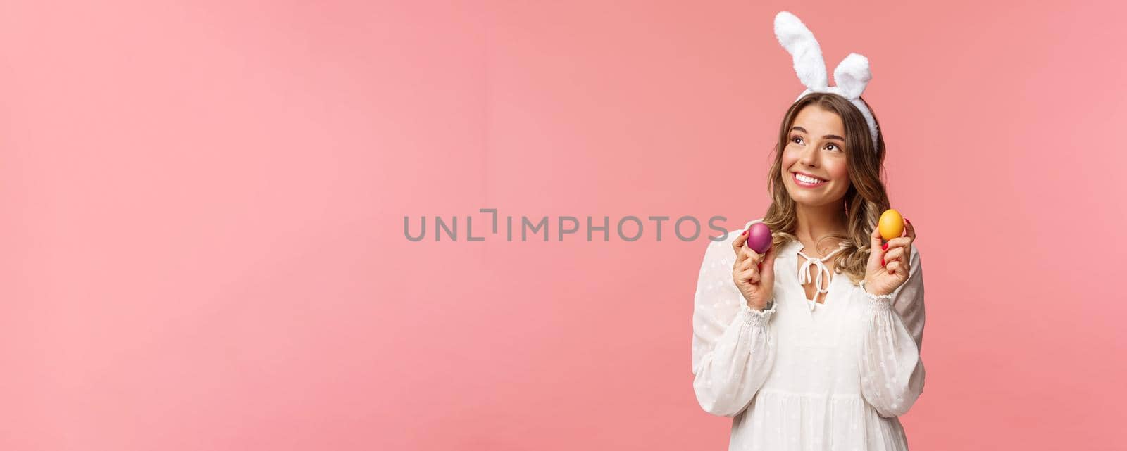 Holidays, spring and party concept. Portrait of dreamy, tender young blond girl feeling happy, celebrating religious day, holy Easter, wear rabbit ears and white dress, hold painted eggs, look away.