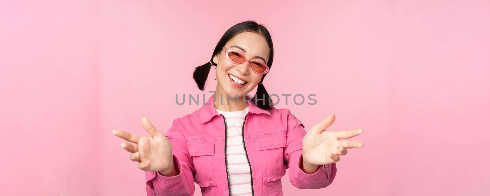 Happy stylish korean girl in sunglasses, reaching hands, receiving, smiling friendly and welcome you, standing over pink background.