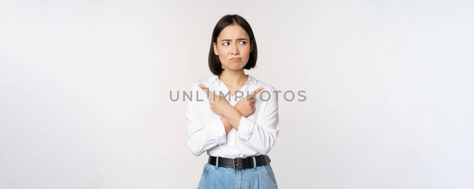 Indecisive asian woman pointing fingers sideways, pointing fingers and looking clueless, confused with choices, standing over white background.