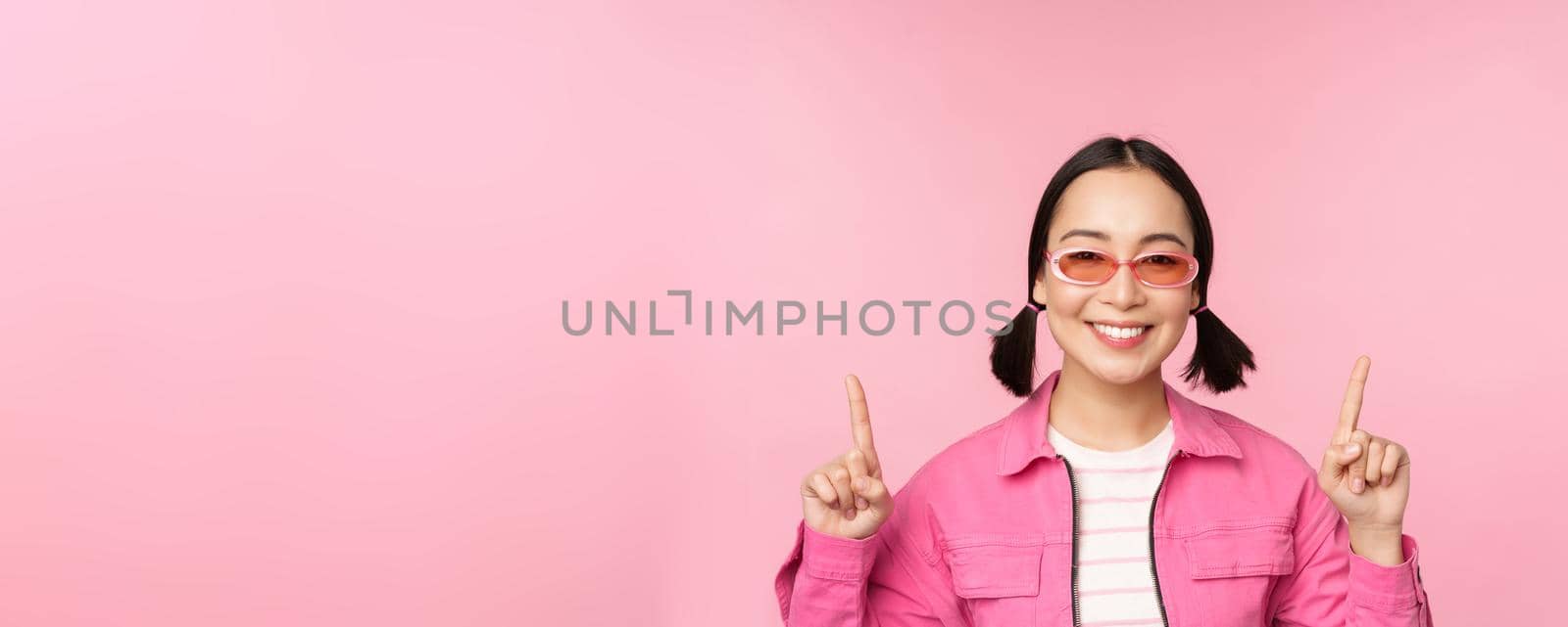 Close up portrait of modern korean female model, wears sunglasses, points fingers up, shows advertisement, promo banner, pink background. Copy space
