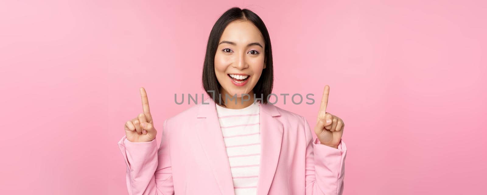 Smiling korean businesswoman, pointing fingers up, showing advertisement, banner or logo on top, standing in suit over pink background.
