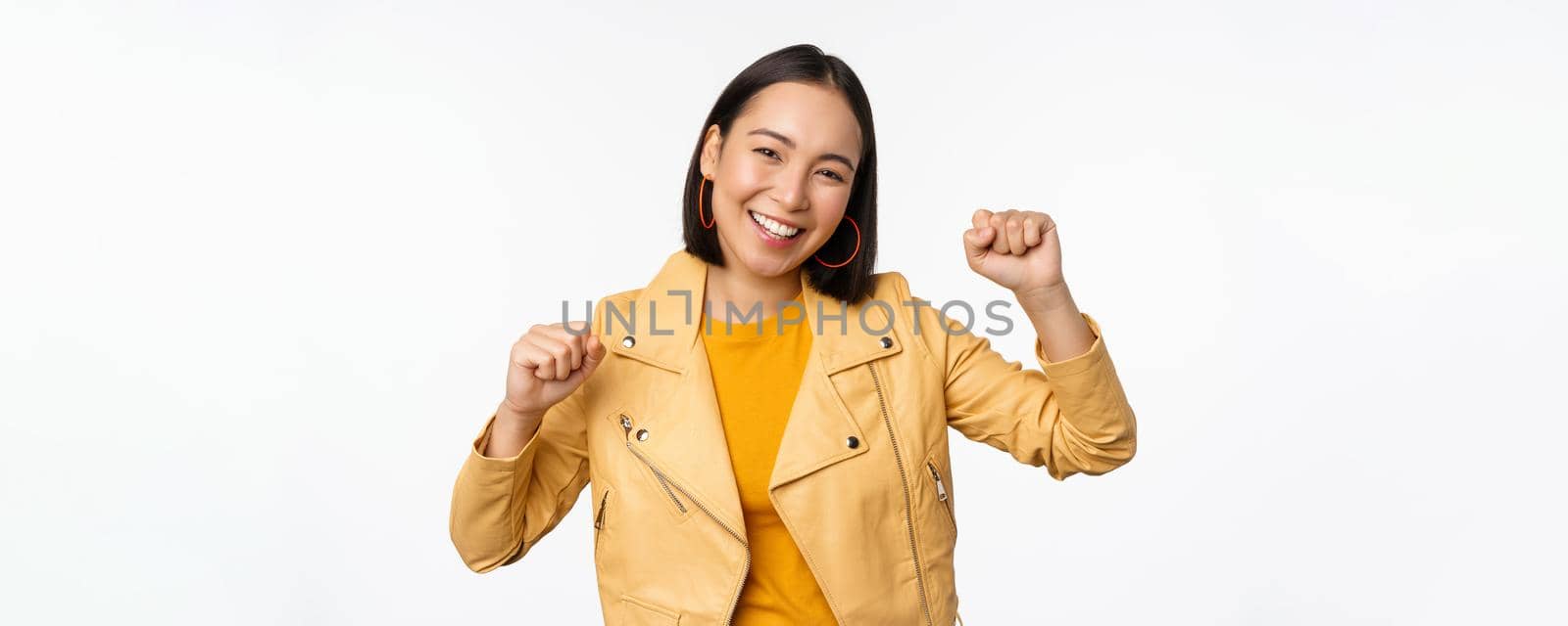 Image of asian woman dancing and smiling happy, celebrating victory, triumphing, standing over white background by Benzoix