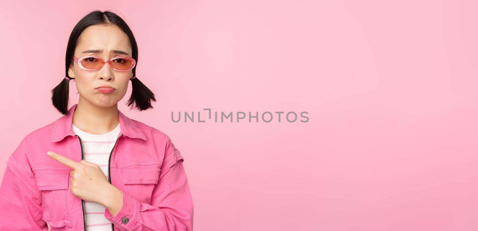 Potrait of cute korean girl in sunglasses, pointing left and looking disappointed, sulking upset, standing over pink background.