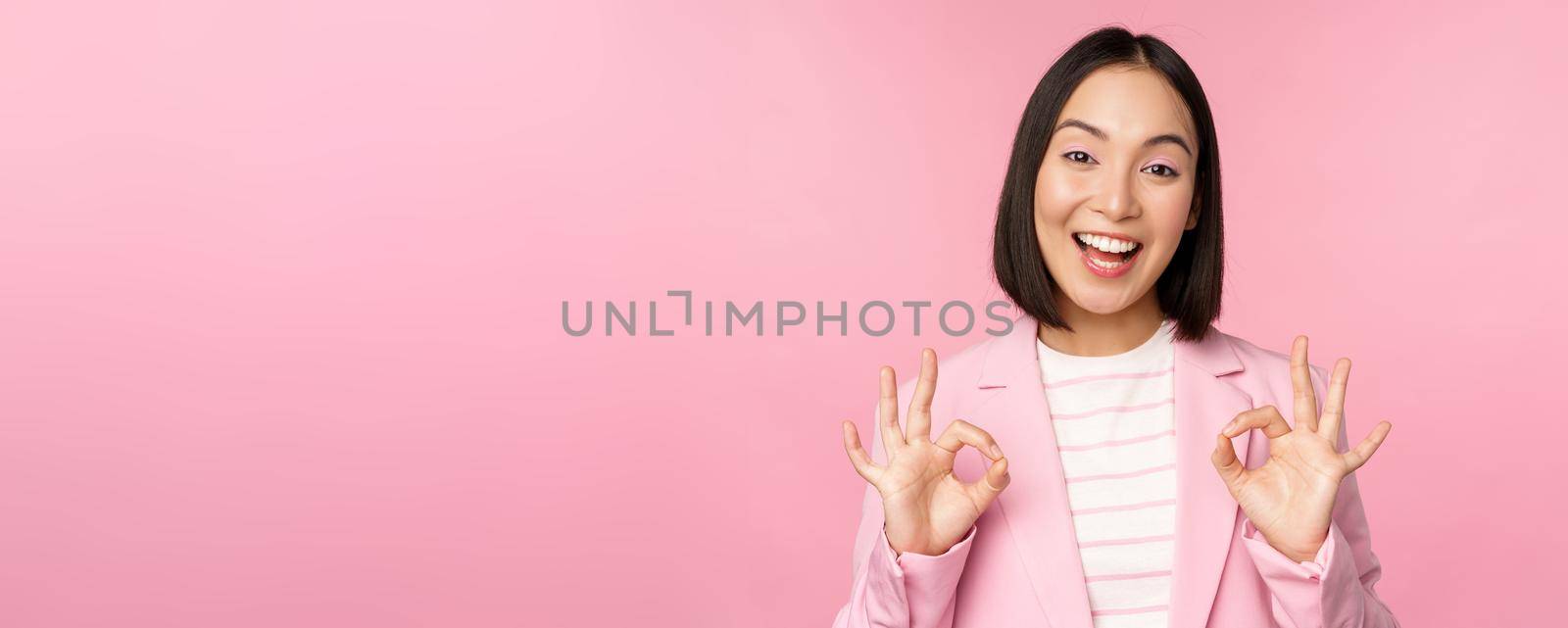 Okay, excellent. Businesswoman in corporate suit, showing ok, approval gesture, recommending smth, give positive feedback and smiling pleased, posing over pink background.