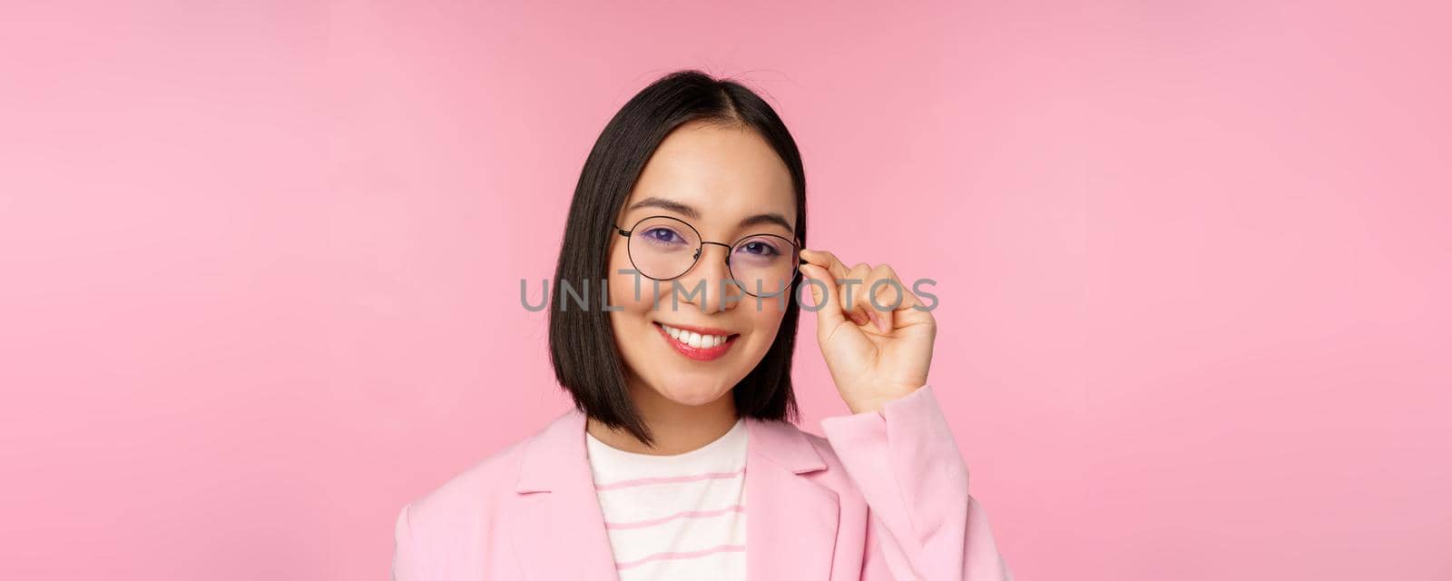 Successful asian businesswoman in glasses, smiling and looking professional at camera, wearing suit, standing over pink background by Benzoix