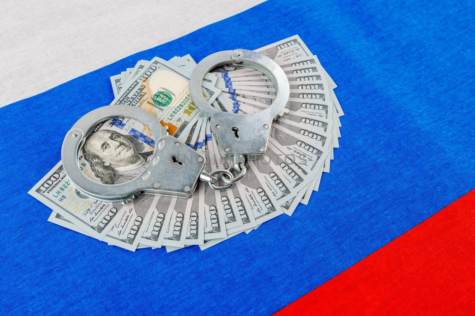 silver metal police handcuffs over paper dollar banknotes of United States of America over national flag of Russian Federation, concept of foreign currency prohibition