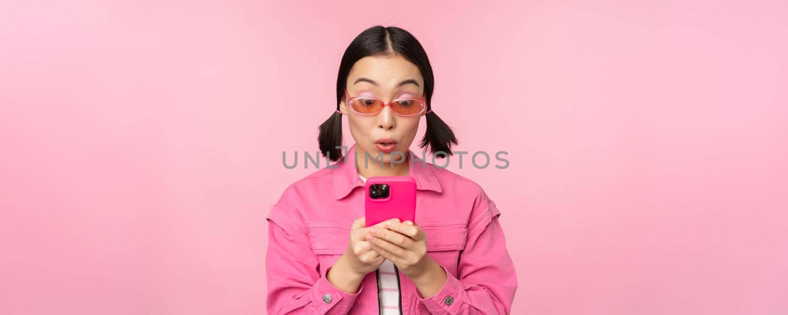 Portrait of asian girl in sunglasses using smartphone. Woman looking at mobile phone, browsing in app, standing over pink background.