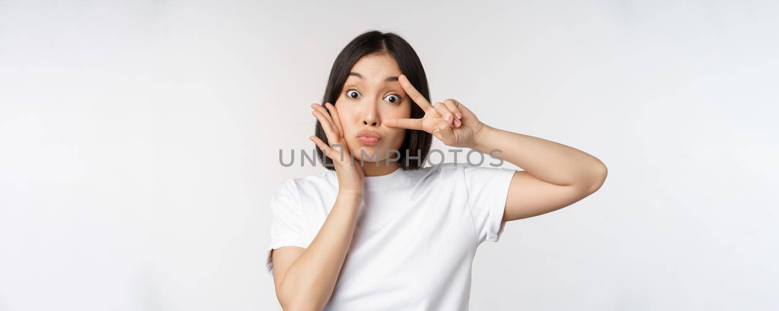 Cute asian girl posing with kawaii v-sign, peace gesture near face, standing in tshirt over white background. Copy space