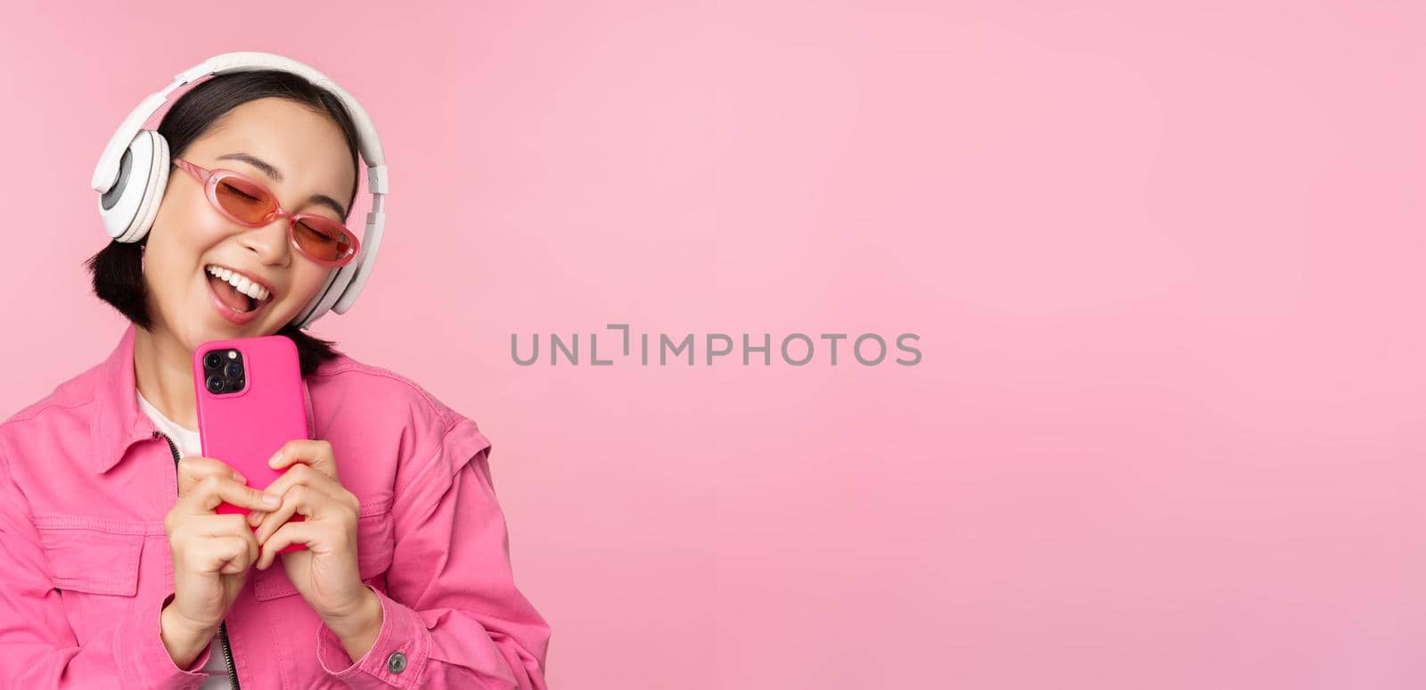 Stylish asian girl in headphones, listening music and taking photos on mobile phone, using smartphone, standing over pink background.