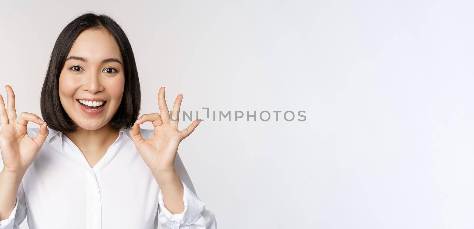 Close up head portrait of asian girl showing okay, ok sign and smiling satisfied, recommending, being pleased, praise and make compliment, white background.