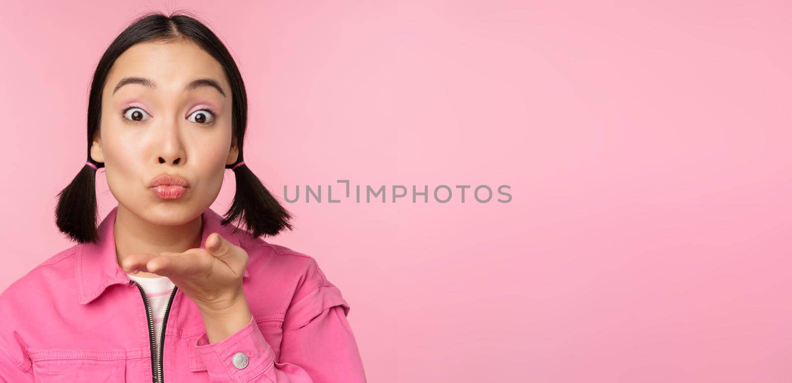 Close up portrait of beautiful silly asian girl, sending air kiss, mwah at camera, blowing, standing over pink background.