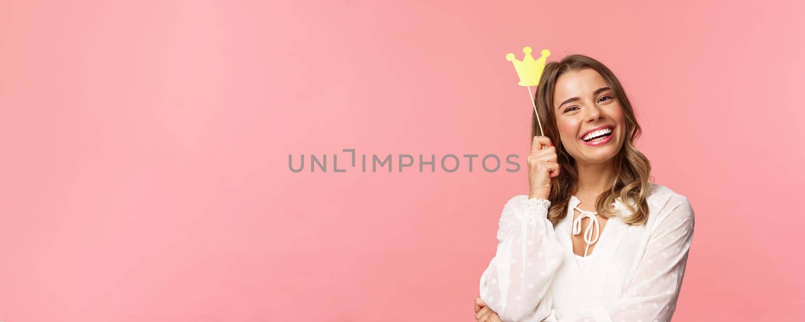 Spring, happiness and celebration concept. Close-up portrait of charming smiling, lovely blond girl holding small queen crown on stick, laughing joyfully, feel empowered and happy, pink background.