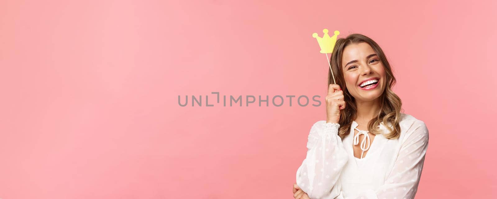 Spring, happiness and celebration concept. Close-up portrait of charming smiling, lovely blond girl holding small queen crown on stick, laughing joyfully, feel empowered and happy, pink background by Benzoix