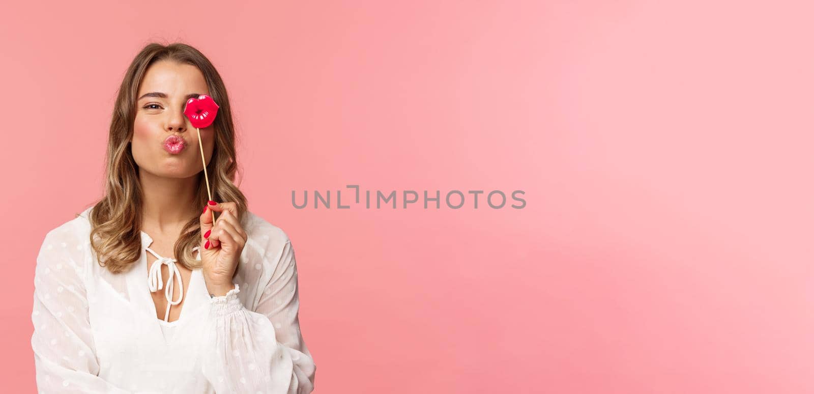 Spring, happiness and celebration concept. Close-up portrait of feminine lovely blond girl in white dress, folding lips in kiss and holding carton stick, partying having fun, stand pink background.