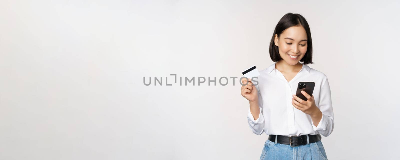 Online shopping concept. Korean woman holding credit card and looking at smartphone app, buying, order delivery in mobile phone application, standing over white background.