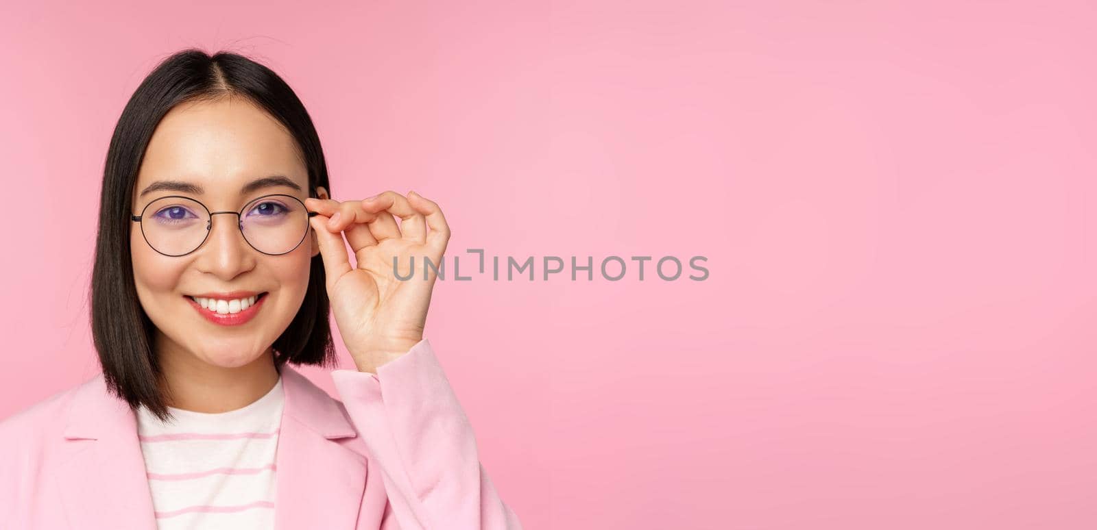 Close up portrait of asian corporate woman, professional businesswoman in glasses, smiling and looking confident at camera, pink background.