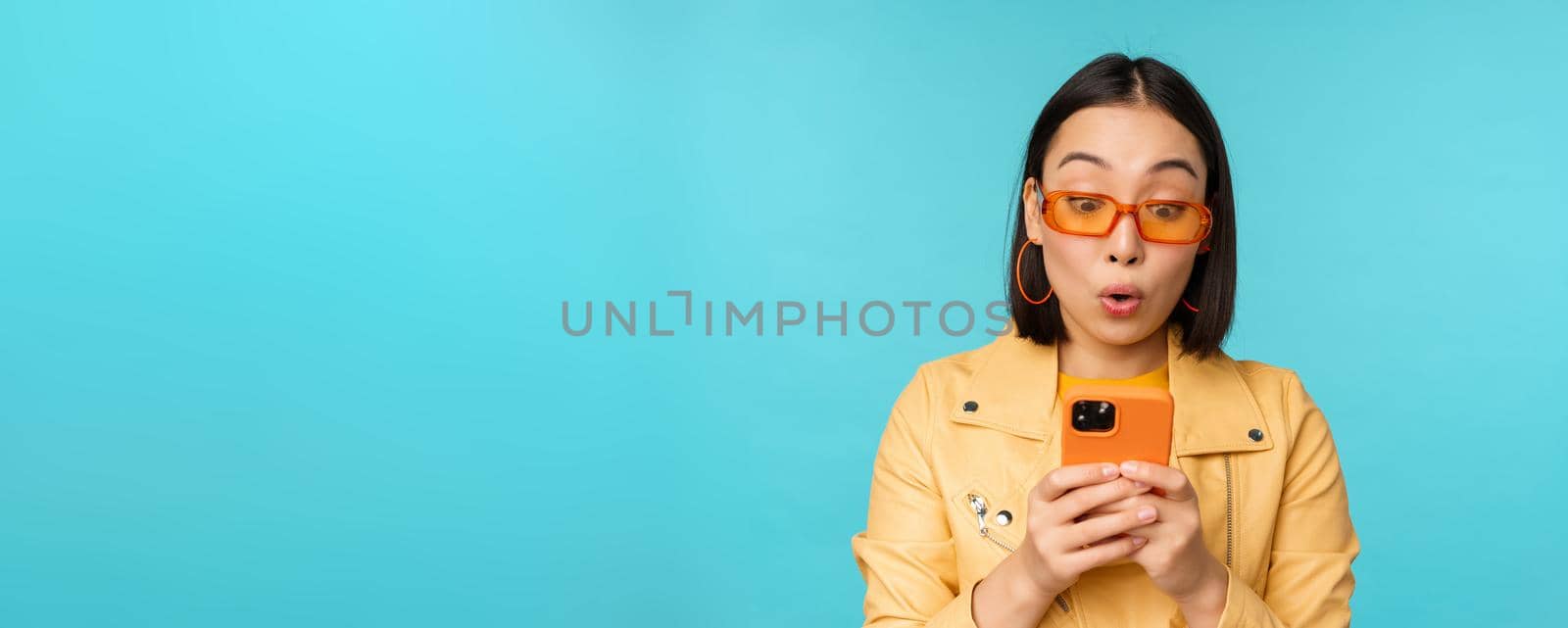 Portrait of asian woman in sunglasses, looking at mobile phone with surprised face expression, standing over blue background. Copy space